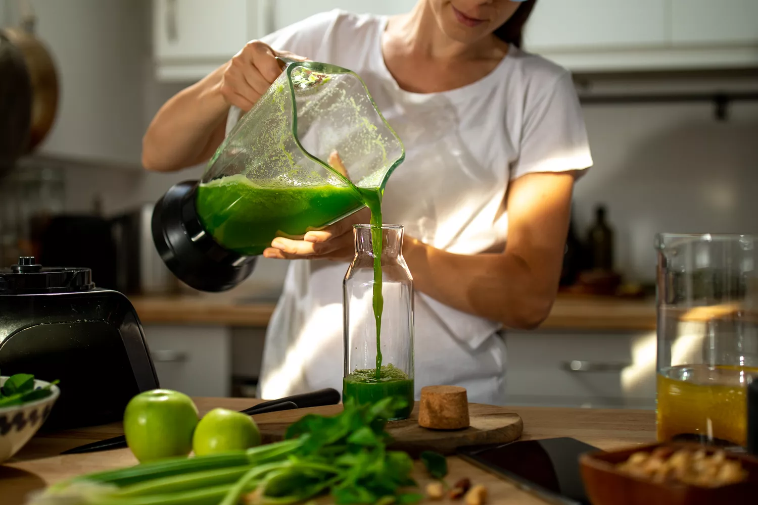 Woman wearing a white t-shirt standing at a kitchen counter pouring a green smoothie into a glass jar. 